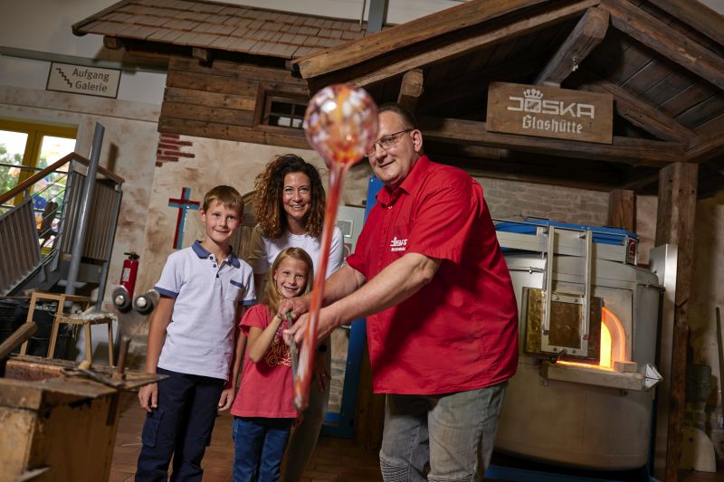 In einer charmanten Glashütte in der Nähe des Hotels Bodenmais zieht ein gelernter Glasbläser eine Familie mit zwei Kindern in seinen Bann und präsentiert unter einem rustikalen Holzschild die Kunst der Glasherstellung.
