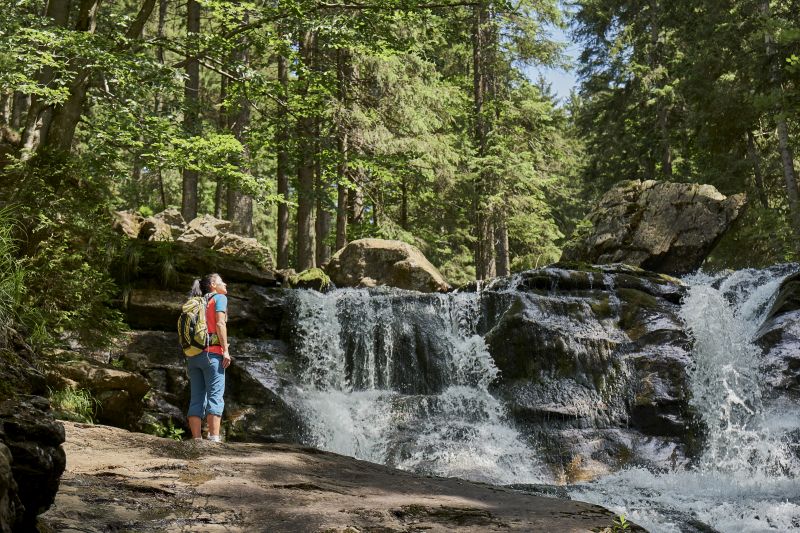 Eine Person mit einem Rucksack steht an einem Wasserfall in einem üppigen Wald in der Nähe von Bodenmais und blickt auf das herabstürzende Wasser.