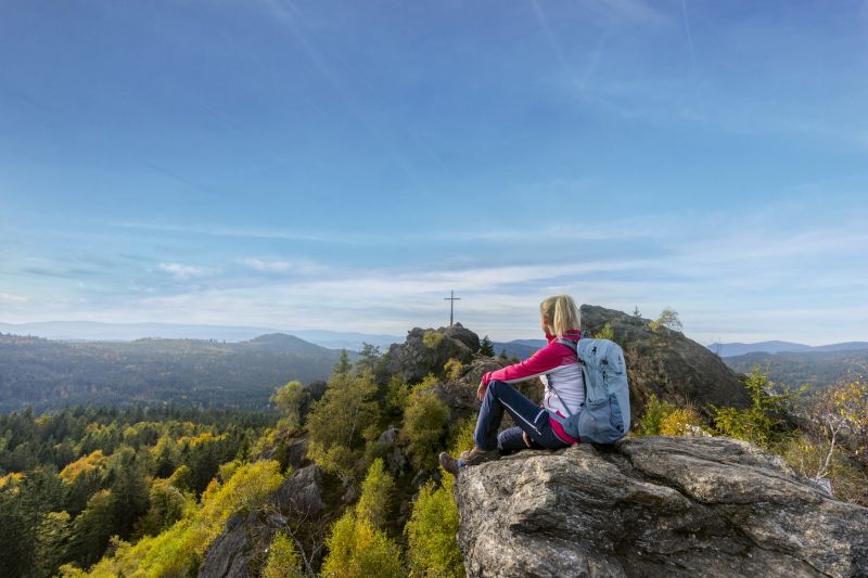 Eine Person mit einem Rucksack sitzt auf einem Felsen und genießt die malerische Bergsicht bei Bodenmais. In der Ferne ist ein Kreuz zu sehen.