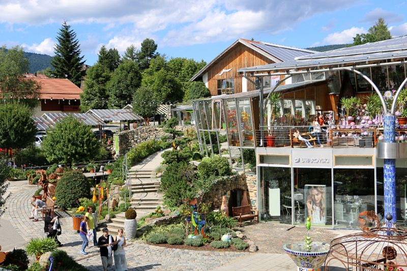 Eine sonnenbeschienene Szene im Freien zeigt das Hotel Bodenmais mit seiner Glasfront, umgeben von üppigem Grün. Menschen schlendern an lebhaften Sitzbereichen vorbei und genießen die lebendige Atmosphäre von Bodenmais an diesem schönen Tag.