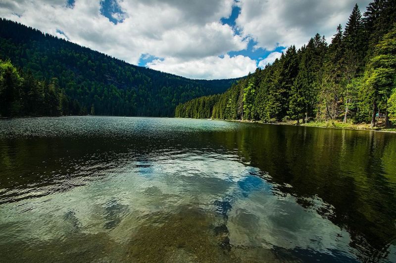 Eingebettet in der Nähe eines klaren Sees, in dem sich die Wolken spiegeln, und umgeben von dichten grünen Wäldern und Bergen unter einem teilweise bewölkten Himmel, bietet das Hotel Bodenmais eine ruhige Zuflucht in die Schönheit der Natur.