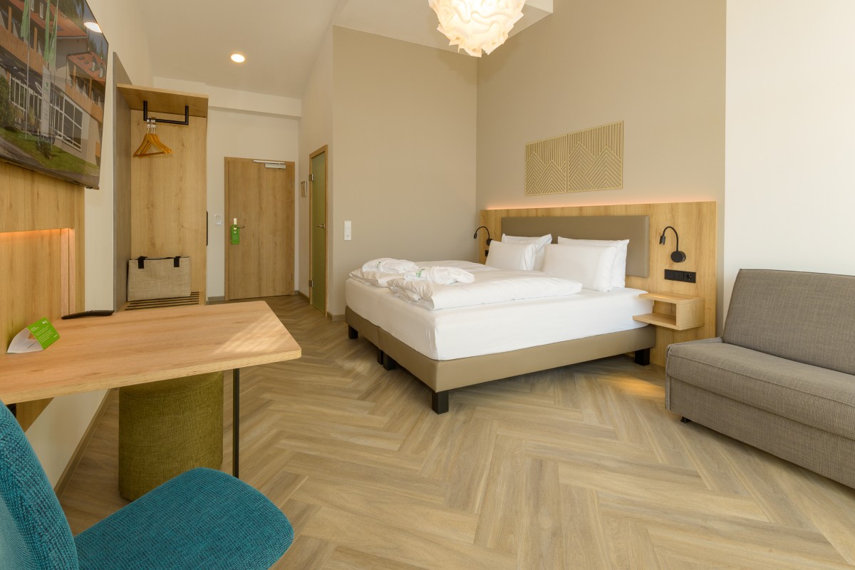 Entdecken Sie den Charme unseres minimalistischen Zimmers im Hotel Bodenmais mit einem gemütlichen Doppelbett, einem weichen Sofa, einem eleganten Schreibtisch und eleganten Holzakzenten. Sanfte Beleuchtung ergänzt den Fischgrätboden und sorgt für eine beruhigende Atmosphäre.