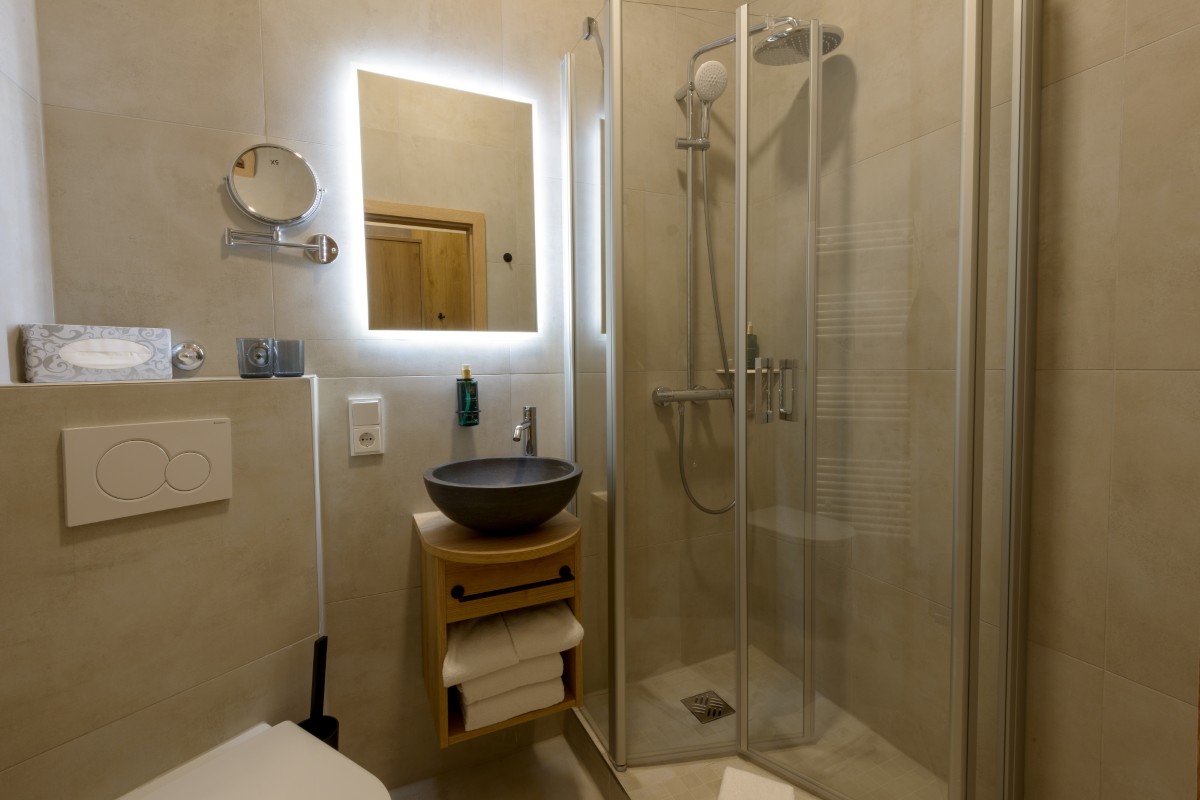 Ein modernes Badezimmer im Hotel Bodenmais verfügt über eine elegante Glasdusche, einen runden Spiegel, einen beleuchteten Kosmetikspiegel, ein Waschbecken und praktische Ablageflächen.