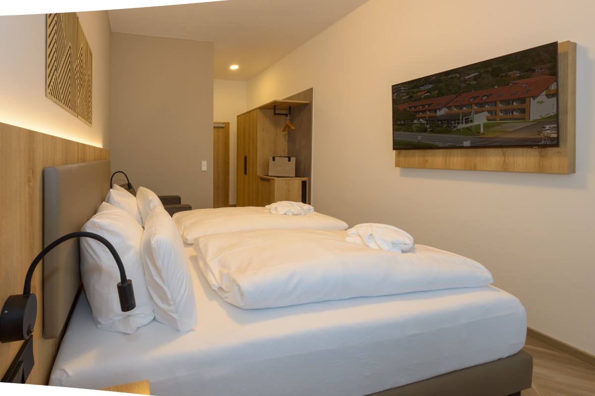 Modernes Zimmer im Hotel Bodenmais mit zwei ordentlich gemachten Betten, Wandfernseher und warmer Beleuchtung. Minimalistisches Dekor und Holzakzente.