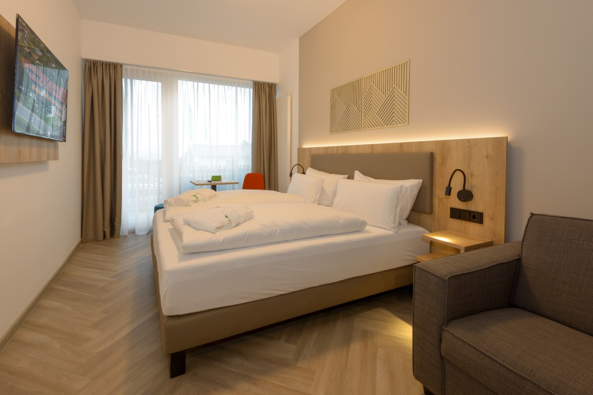 Erleben Sie ein modernes Zimmer im Hotel Bodenmais mit einem geräumigen Bett, einem an der Wand montierten Fernseher, einem gemütlichen Sessel und einem mit transparenten Vorhängen geschmückten Fenster.