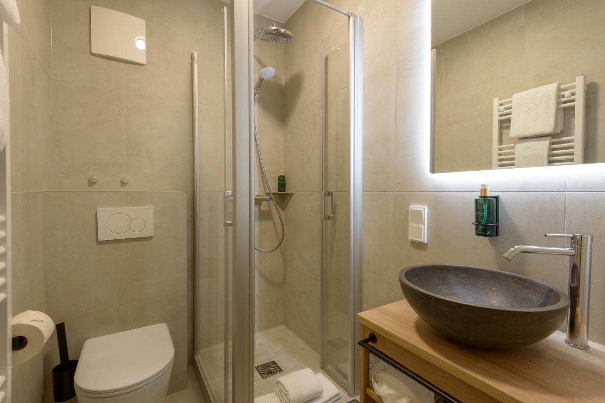 Erleben Sie den Charme eines modernen Badezimmers im Hotel Bodenmais mit einer Glasdusche, einem runden Steinwaschbecken auf einer Holzplatte, einer Toilette und einem großen Spiegel.