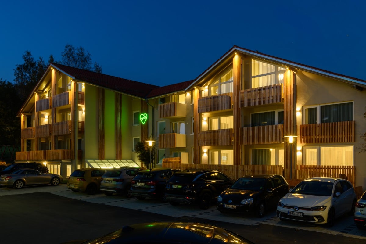 Ein modernes, warm beleuchtetes Mehrfamilienhaus bei Nacht mit parkenden Autos davor und einer grünen Leuchtreklame an der Fassade, die an das stylische Hotel Bodenmais erinnert.