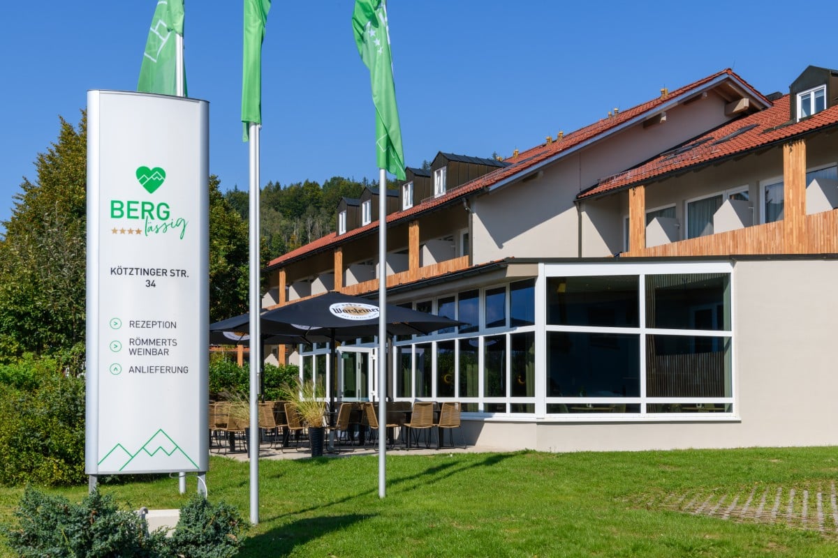 Die Außenfassade des Gebäudes erinnert an das Hotel Bodenmais, mit einem Schild mit der Aufschrift „BERG 34“ und gemütlichen Sitzgelegenheiten im Freien unter Sonnenschirmen. Drei grüne Fahnen flattern im Wind und laden die Gäste ein, den Charme von Bodenmais zu erleben.