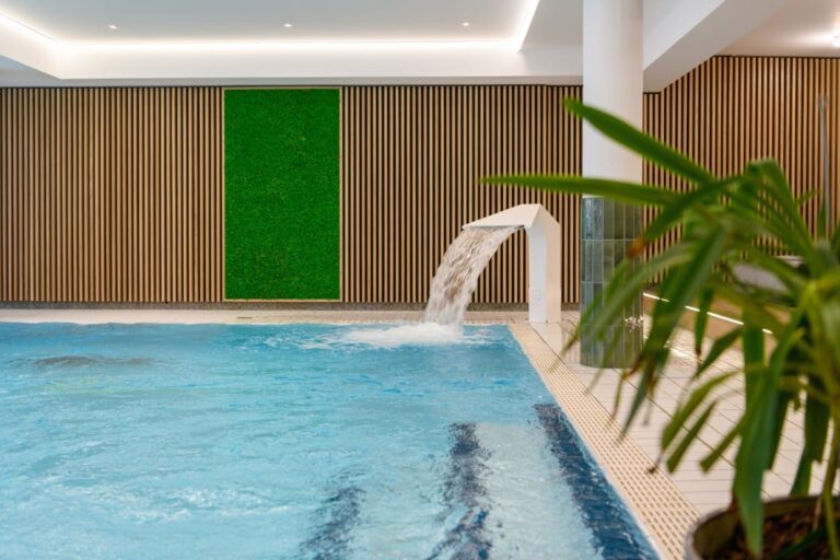 Erleben Sie Ruhe im Hotel Bodenmais mit seinem Innenpool mit modernem Wasserspiel, ergänzt durch elegante Holzpaneelwände und einer großen Grünpflanze im Vordergrund.