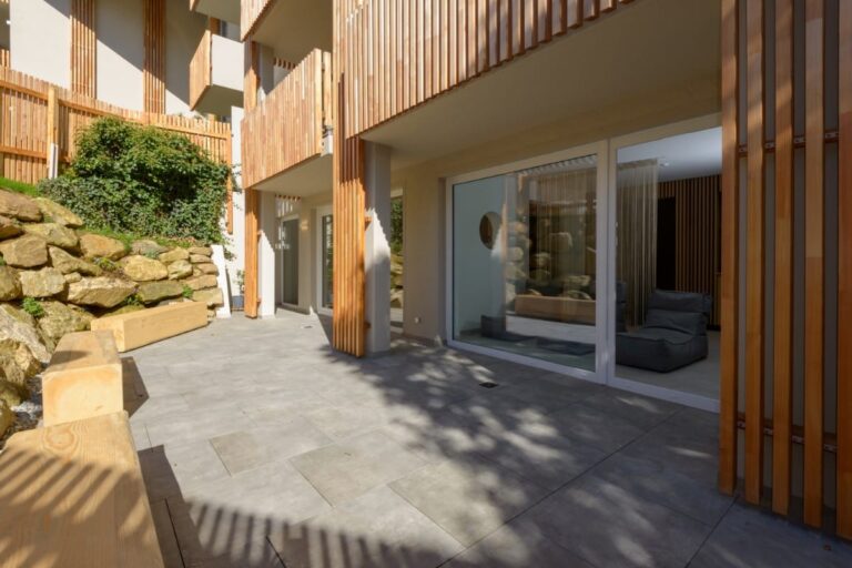 Moderne Terrasse mit Holzakzenten, großen Fenstern und Steinmauern. Sonnenlicht wirft Schatten auf den Steinfliesenboden und schafft eine warme und einladende Atmosphäre, die an das elegante Design des Hotels Bodenmais erinnert.