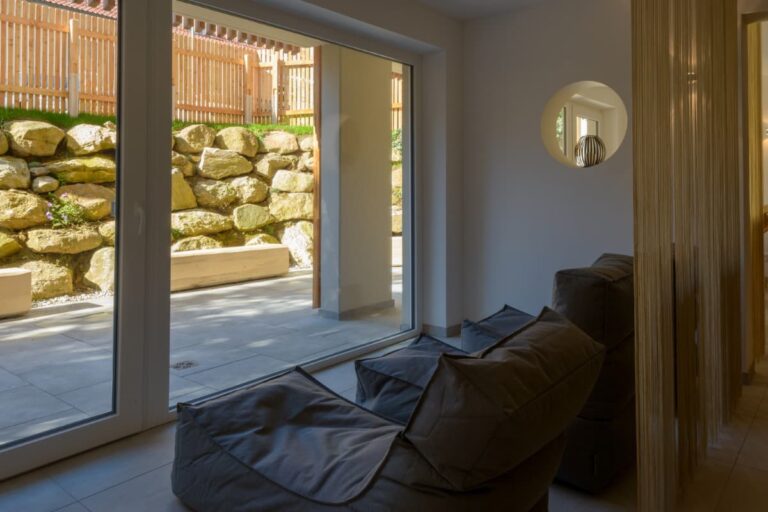 Zwei bequeme Stühle zieren einen modernen Raum mit großen Fenstern und bieten einen herrlichen Blick auf eine Steinmauer und einen Holzzaun. Erleben Sie diese Ruhe im Hotel Bodenmais, wo Komfort auf malerische Eleganz trifft.