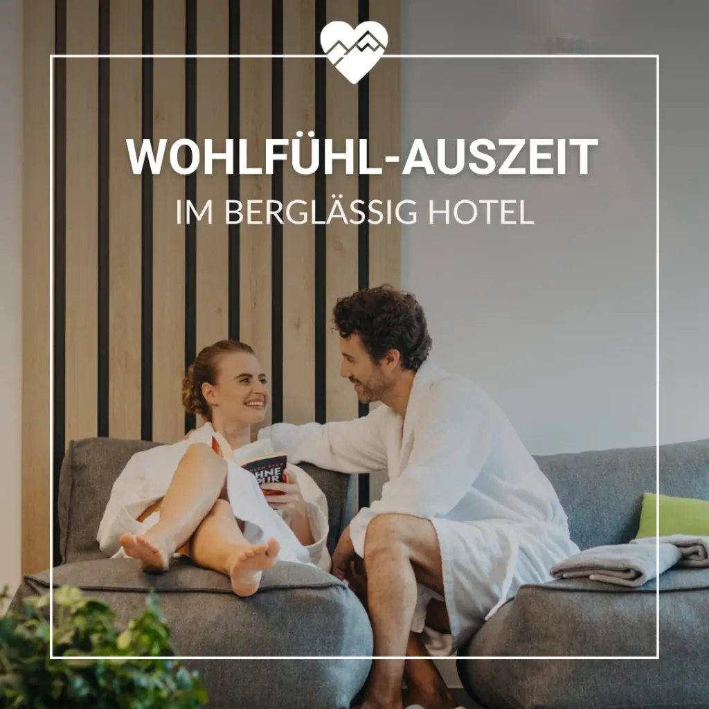 Ein l&auml;chelndes Paar in wei&szlig;en Gew&auml;ndern entspannt sich auf einem Sofa in einem modernen Hotel, dar&uuml;ber der Text "Wohlf&uuml;hl-Auszeit".