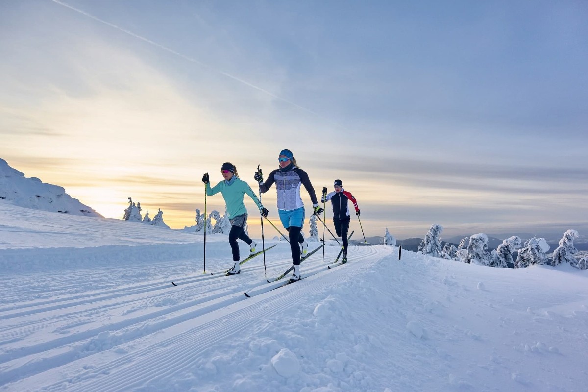 Drei Personen beim Skilanglauf auf einer verschneiten Loipe in einer malerischen Winterlandschaft bei Sonnenuntergang.