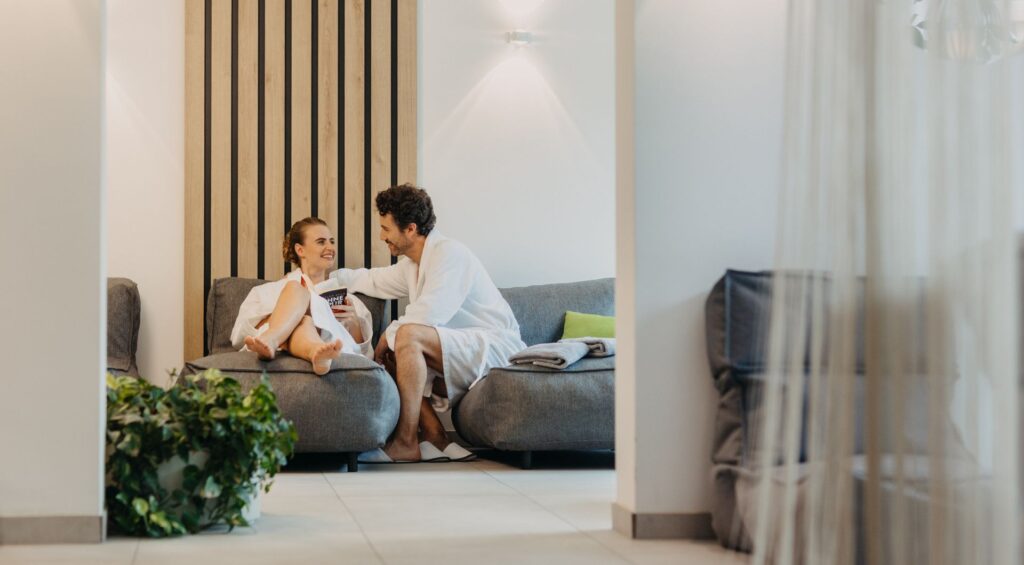 Ein Paar in wei&szlig;en Badem&auml;nteln entspannt sich und l&auml;chelt einander an, w&auml;hrend es auf Sofas in einem modernen, hellen Wohnzimmer sitzt.